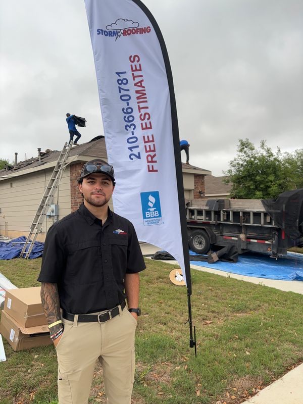 Man stands in front of a roofing service sign with workers on a roof in the background.