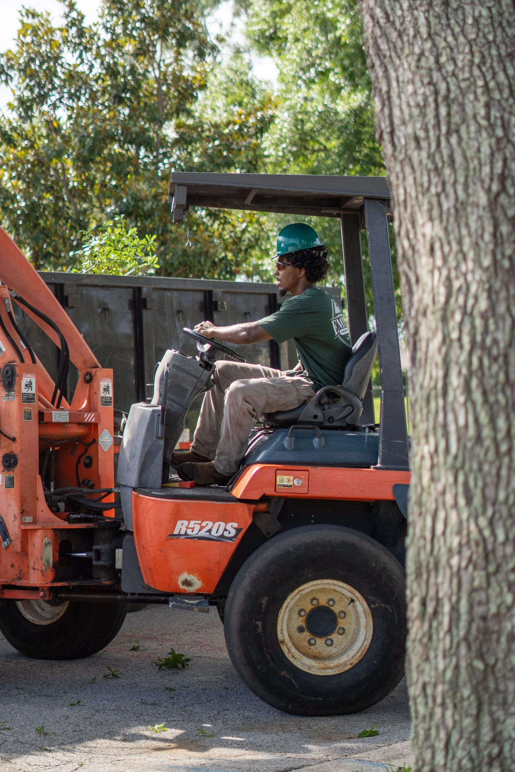 Worker operating an orange forklift outdoors wearing a green hard hat.