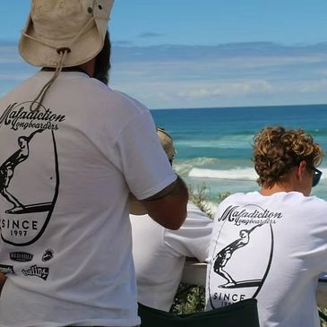 Two men wearing matching white Maladiction Longboards t-shirts by the ocean.