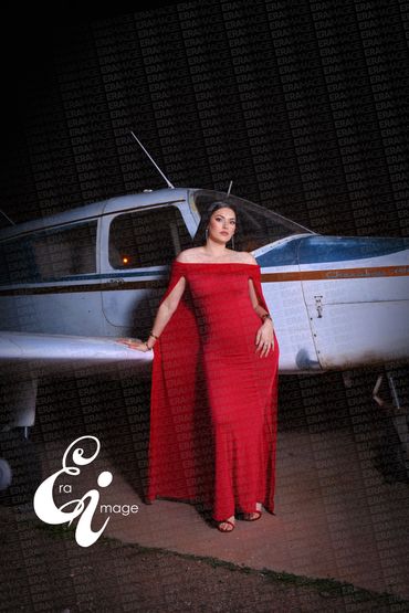 Woman in elegant red dress posing beside a small airplane at night.