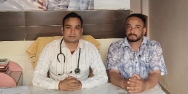 Two men seated at a desk in a healthcare setting with a medical banner behind them.