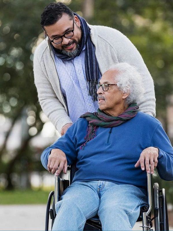 Man pushing elderly man in wheelchair outdoors, sharing a warm moment.