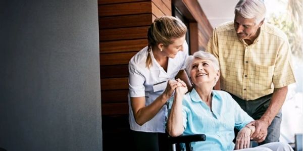 Elderly woman in wheelchair with caregiver and elderly man supporting her.