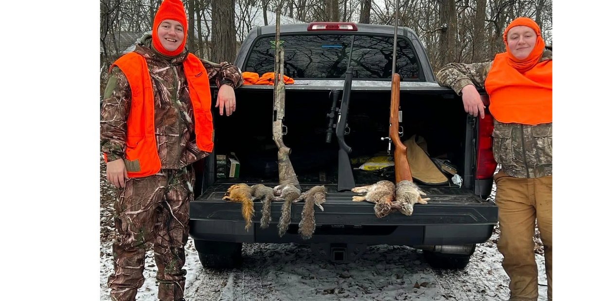 Two hunters in orange gear stand by a truck with hunted squirrels and rifles displayed.
