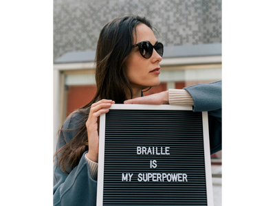 I young woman in sunglasses holding a sign that says "Braille is my superpower"