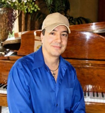 Man in blue shirt and beige cap sitting by a piano.
