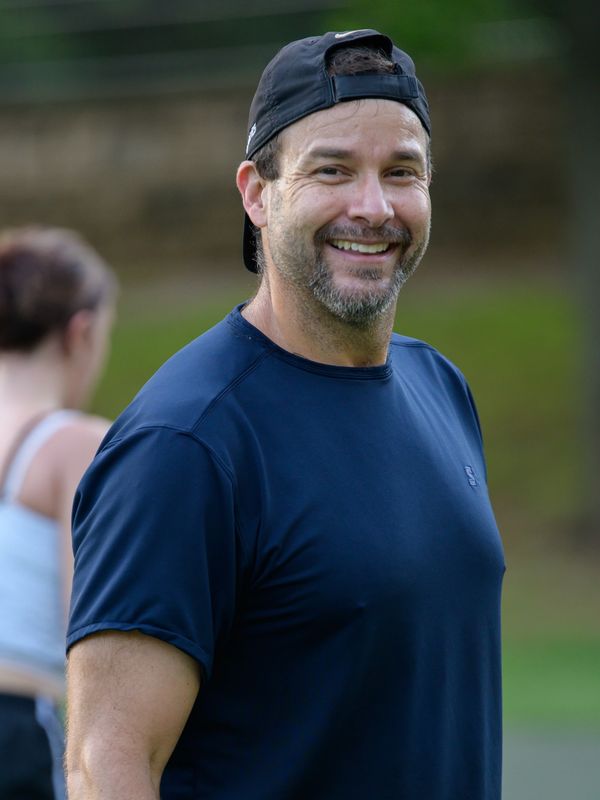 Smiling man in sportswear outdoors with a woman blurred in the background.