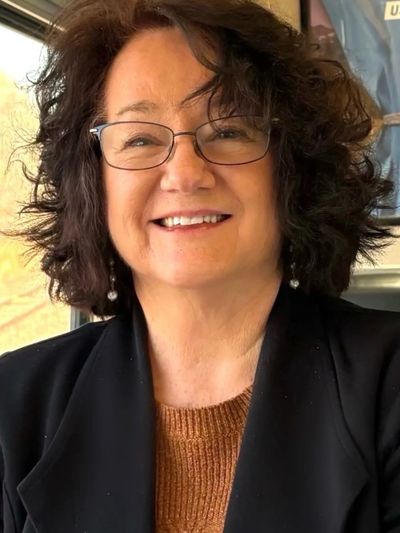 Smiling woman with curly hair and glasses wearing a black jacket.