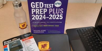Study essentials including GED prep book, calculator, laptop, and water bottle on desk.