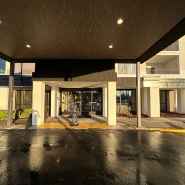 Entrance of a building with a covered walkway and wet pavement reflecting sunlight.