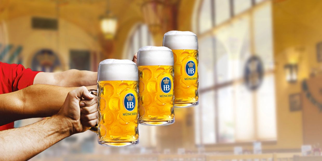 Three hands holding large HB München beer mugs with foam in a warm pub setting.