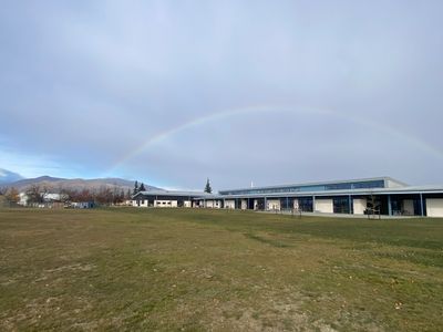 A faint rainbow arches over a modern building with a large grassy field in front.
