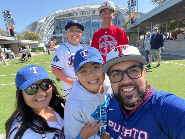 Happy family wearing Texas Rangers gear outside AT&amp;T Stadium on a sunny day.