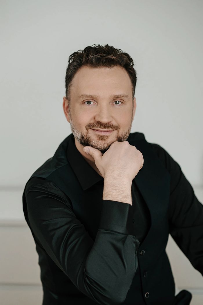 Man with beard wearing black shirt and vest, smiling gently.
