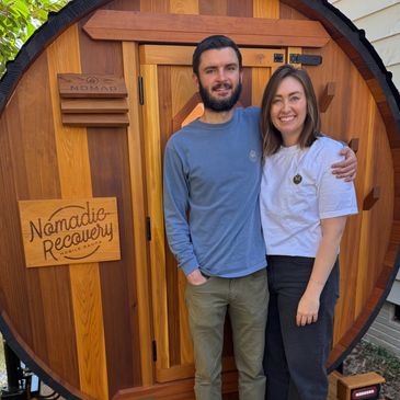 A smiling couple stands in front of a wooden door with 'Nomadic Recovery' sign.