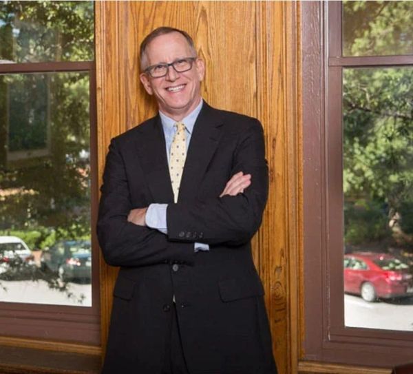 Smiling man in a suit stands with arms crossed by windows.