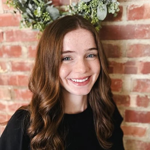 Young woman with long brown hair smiling in front of a brick wall.