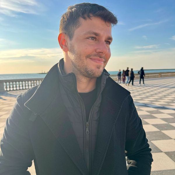 Uomo sorridente con giacca nera al tramonto sul lungomare.