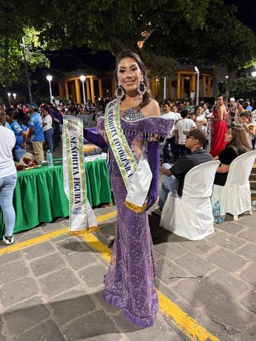Woman in a purple gown with beauty pageant sashes at an outdoor event at night.