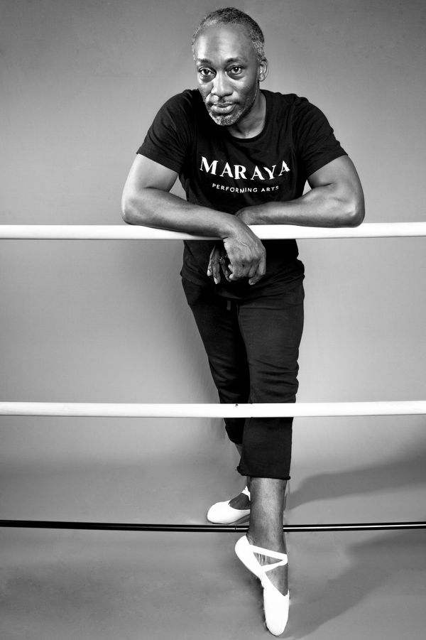 Man in ballet shoes leaning on ballet barre, wearing a Maraya Performing Arts shirt.