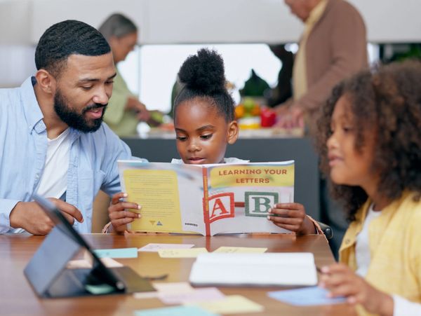 A man helps a young girl read a book titled "Learning Your Letters" while another child sits nearby.