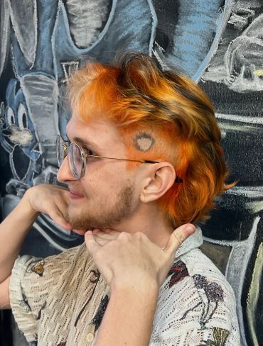 Person with orange hair and glasses posing against graffiti wall.