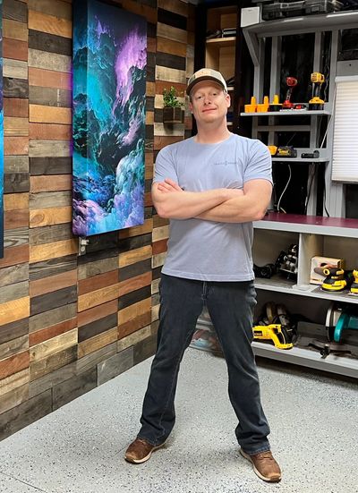 Man standing with crossed arms in a workshop with colorful artwork on the wall.