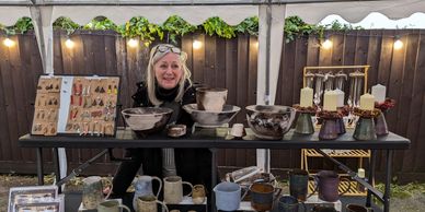 A woman showcases handmade pottery and candles at an outdoor market stall.