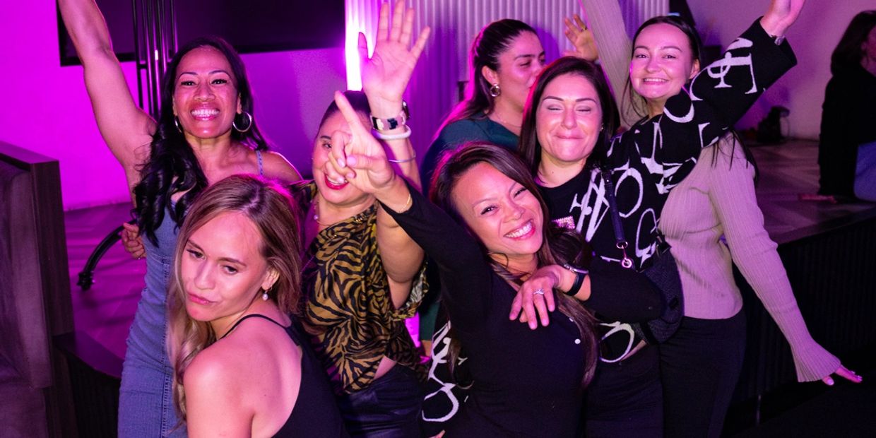 Group of women joyfully posing and dancing under pink party lights.