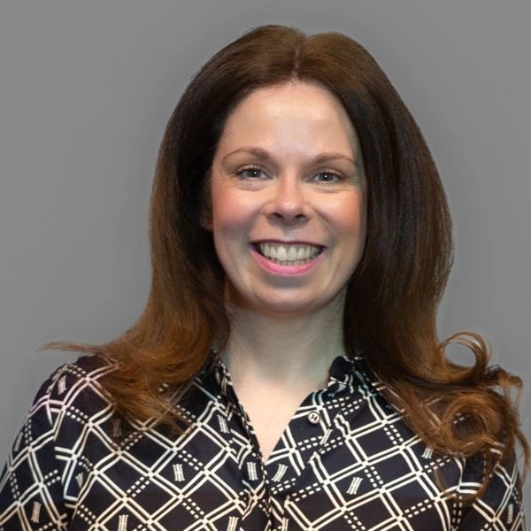 Smiling woman with long brown hair wearing a patterned blouse against a gray background.