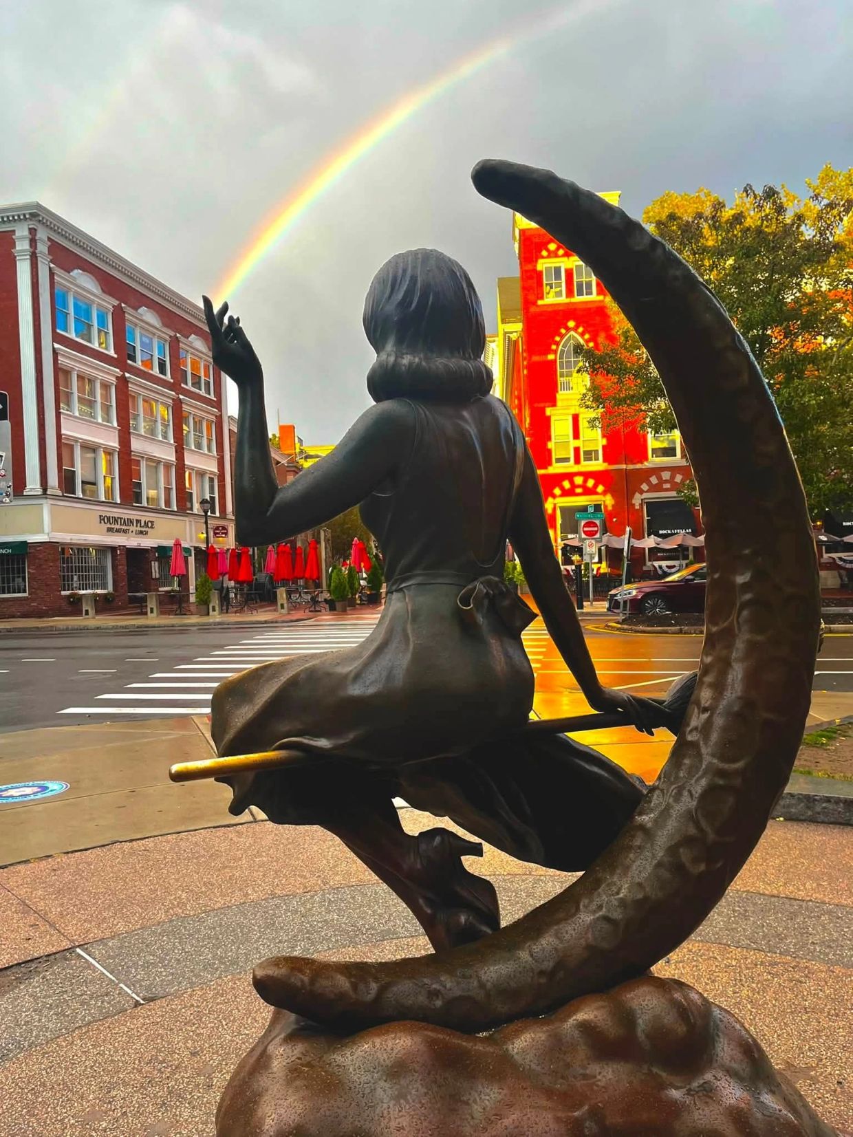 Statue of a girl on a broom with a rainbow in the background.