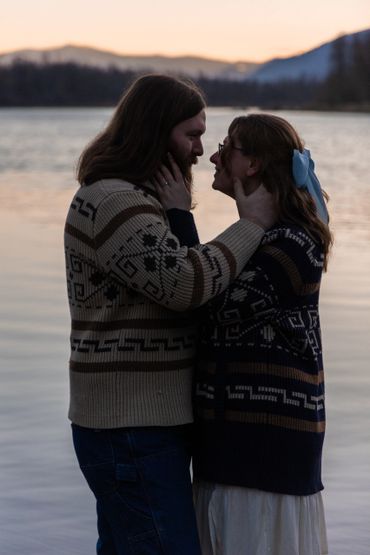 Couple in patterned sweaters sharing a tender moment by the lake at sunset.