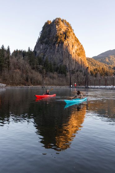Two people kayaking on calm water near a sunlit rocky mountain.
