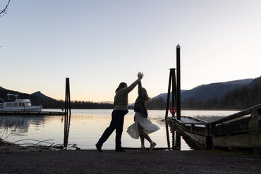 A couple dances by a lakeside at sunset with mountains in the background.
