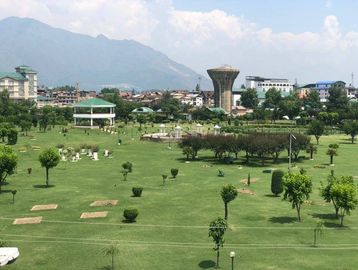 A large green park with trees, gazebos, and buildings in the background under a cloudy sky.