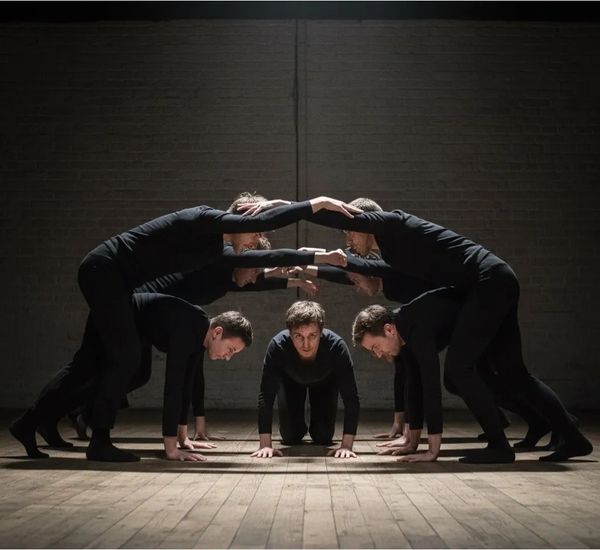 A group of performers in black creating a human arch formation on a wooden floor.