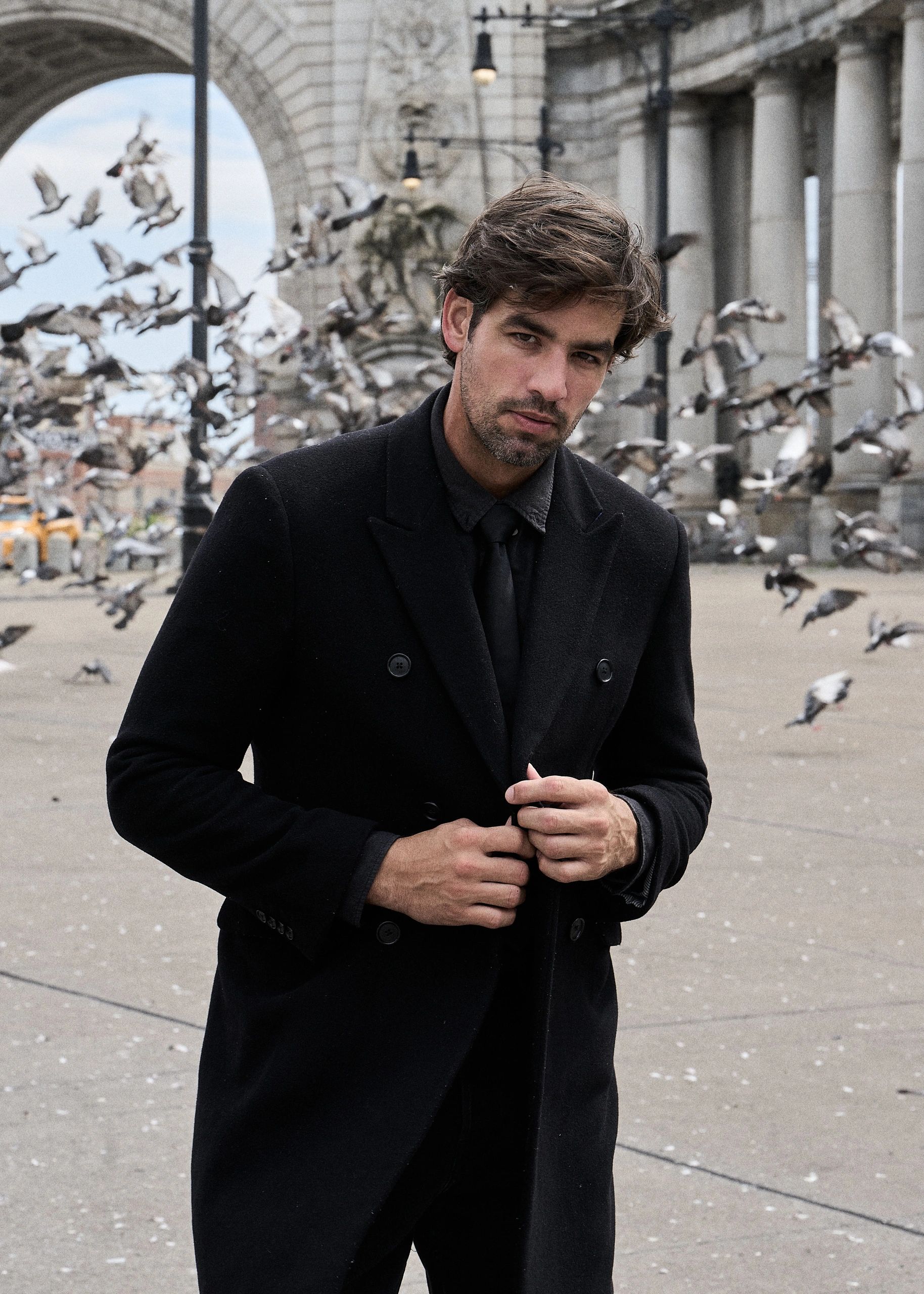 Hombre bien vestido con abrigo negro y palomas volando.