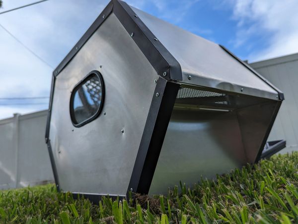 A metallic, geometric structure on grass with a small window and mesh opening.