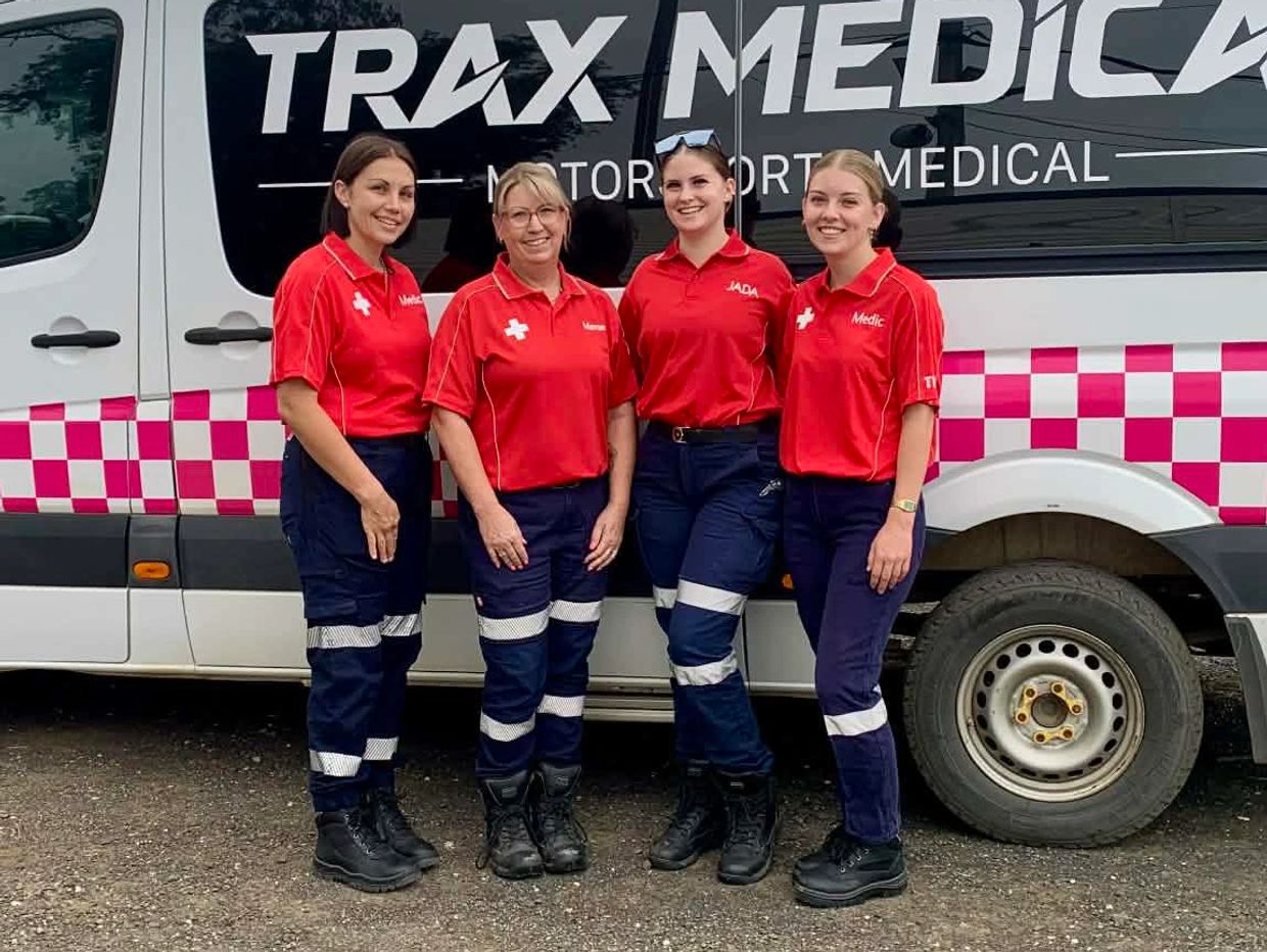 Four female paramedics in red and navy uniforms stand smiling by a medical van.