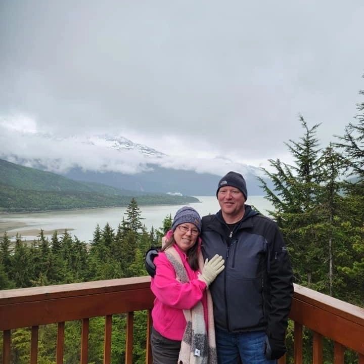Couple dressed warmly, posing on a wooden deck with a scenic mountainous lake backdrop.
