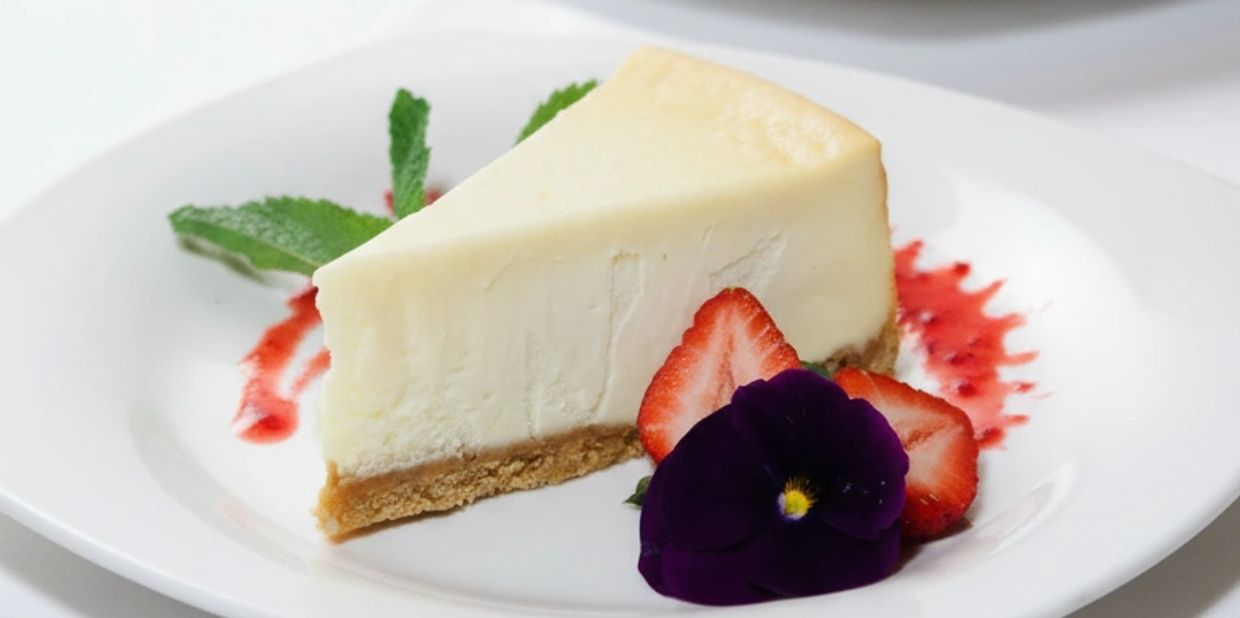 Slice of cheesecake garnished with strawberries and a purple flower on a white plate.