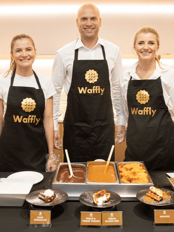 Three smiling staff at a waffle bar wearing black Waffly aprons.