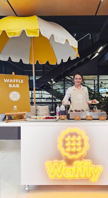 Smiling woman serving waffles at a bright waffle bar with yellow umbrella.
