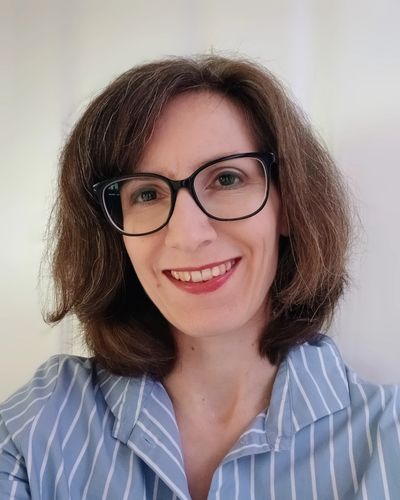 A Caucasian woman with mid-length brown hair and glasses, smiling with a welcoming expression.