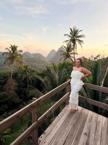 Woman in white dress poses on wooden deck with tropical mountains.