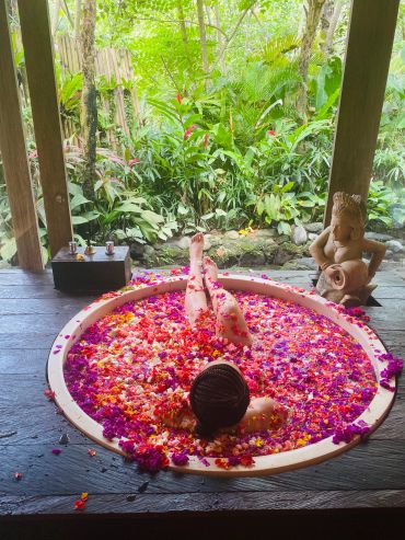 Person relaxing in flower petal-filled tub outdoors.