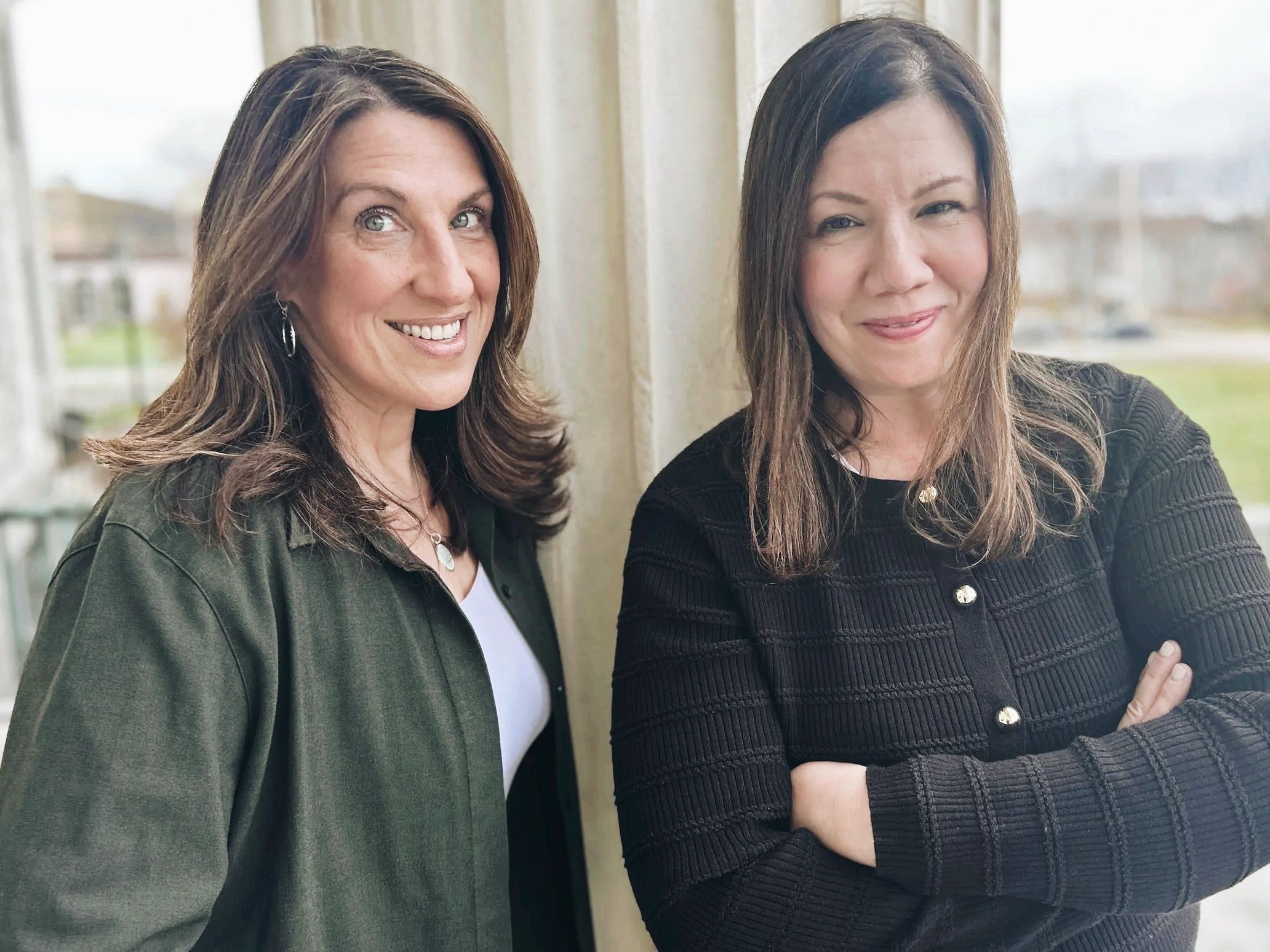 Two women smiling indoors with a bright window background.