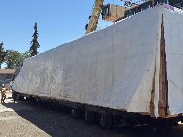 Large modular building section covered in white tarp being transported on a multi-axle trailer.