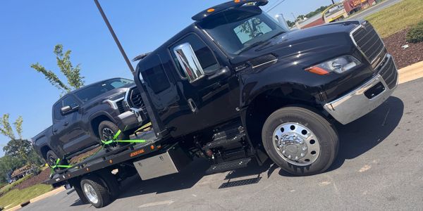 Black flatbed tow truck carrying a gray pickup truck on a sunny day.