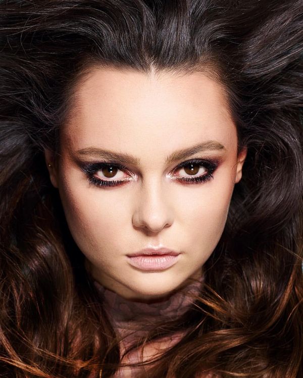 Portrait of a woman with intense smokey eye makeup and voluminous dark hair flowing around her face. Her gaze is direct and captivating, with eyeliner and eyeshadow artfully applied to accentuate her eyes, complemented by a neutral lip color. 